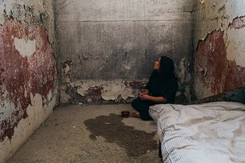 Mulher presa em uma cela, agachada no canto, olhando para a parede